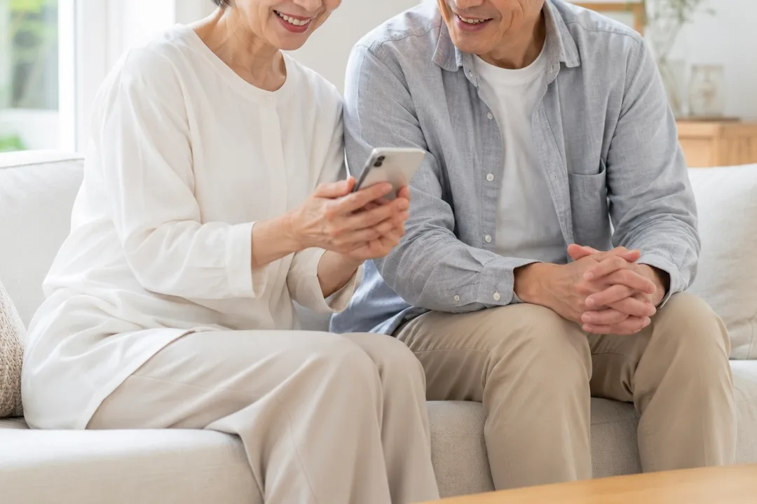 ソファに並んでスマートフォンの画面を一緒に見るシニアの夫婦